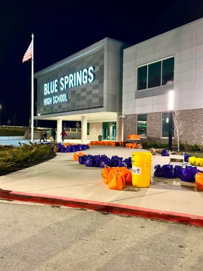 Rows of filled purple and orange donation bags and a Many Parts, One Body barrel staged outside Blue Springs High School at night, ready for distribution